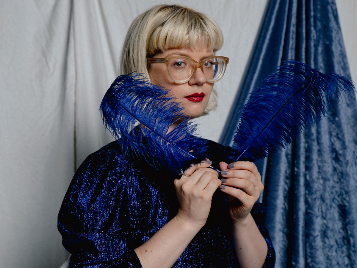 Ein Portrait der Musikerin Mary Ocher, die in einem dunkelblauen Oberteil vor einem grauen Hintergrund sitzt und zwei blaue Federn in beiden H&auml;nden vor sich h&auml;lt.
