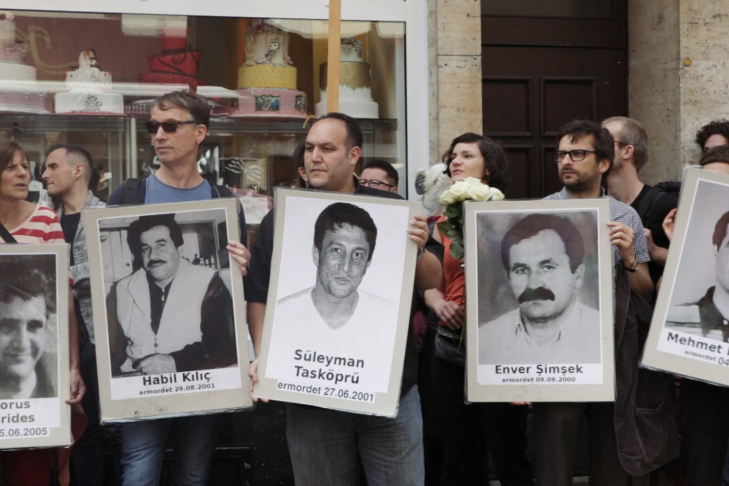 Szene aus dem Film "Der zweite Anschlag", in der man eine Gruppe von Menschen sieht mit Schildern, die an Ermordete erinnern.