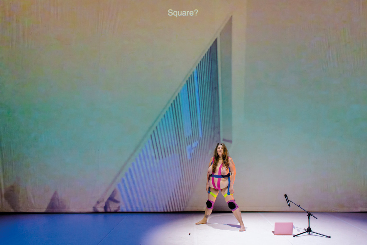 Katy Baird steht halbnackt und mit auseinander gespreizten Beinen vor einer Leinwand-Projektion, schr&auml;g vor ihr stehen ein Mikrofon und ein Laptop auf dem Boden.