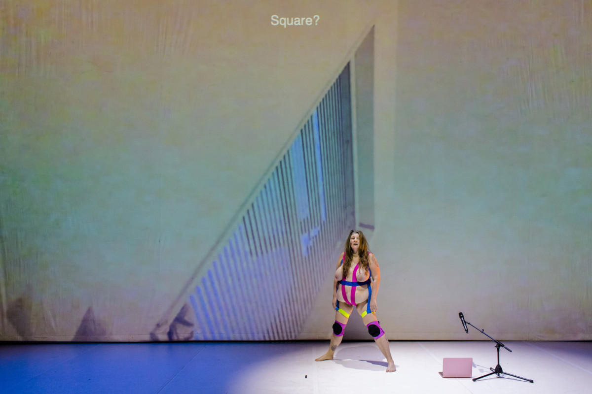 Katy Baird steht breitbeinig vor einer Leinwand, rechts neben ihr stehen ein Laptop und ein Mikrofon auf dem Boden. Sie ist halbnackt und nur mit buntem Klebetape bedeckt.
