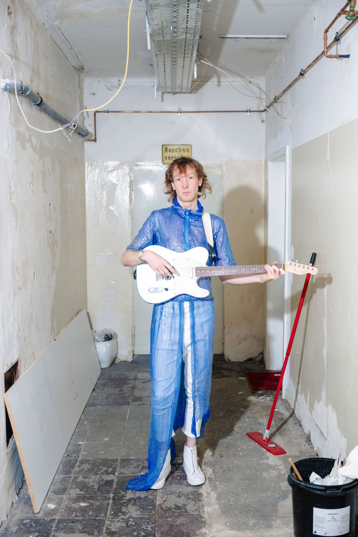 TCHAIR mit wei&szlig;er Gitarre und blauem Pailetteneinteiler in einer Abstellkammer