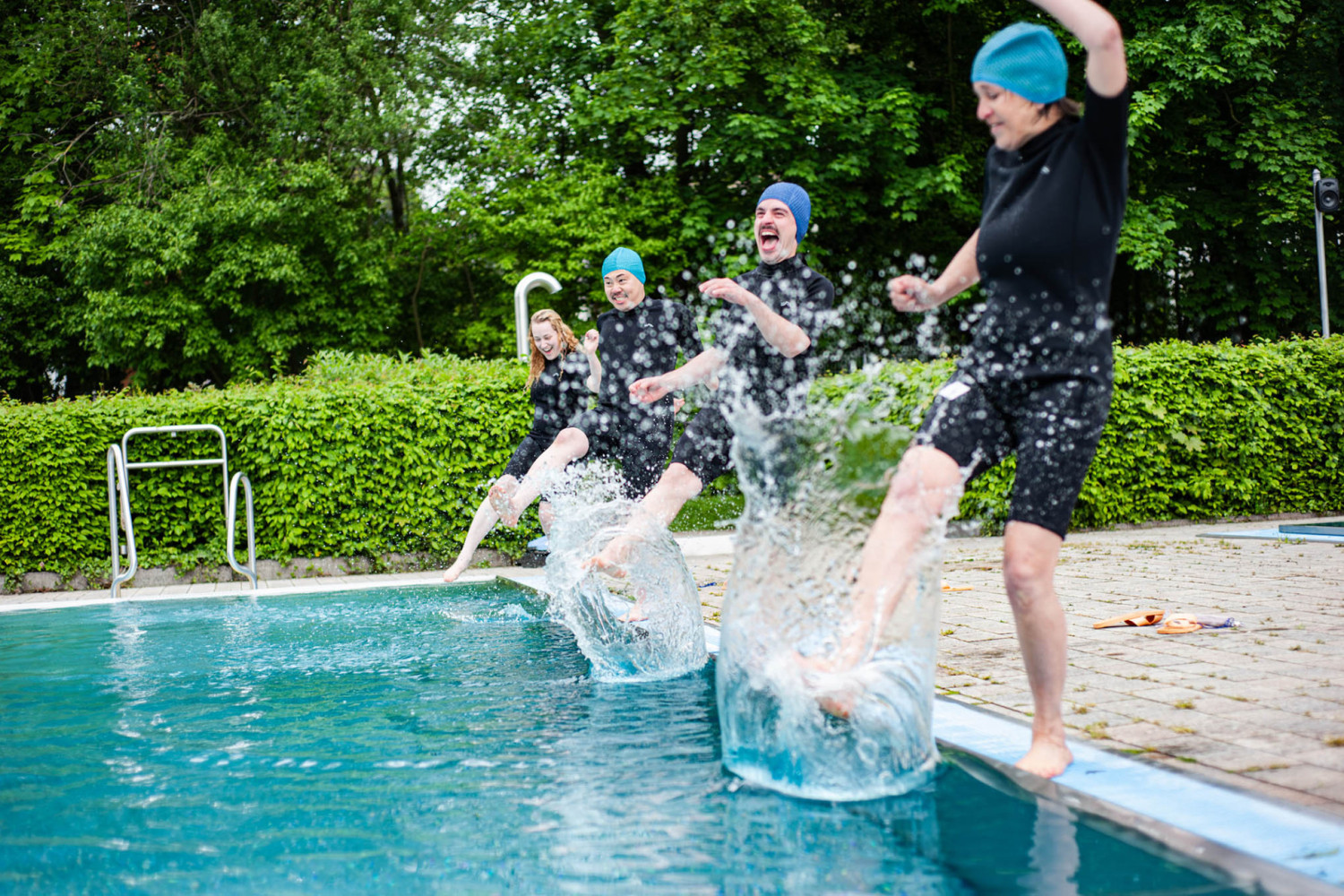 4 Personen stehen mit Badekappe und Schwimmanz&uuml;gen am Rand des Beckens und schlagen mit einem Fu&szlig; auf die Wasseroberfl&auml;che, dass es spritzt.