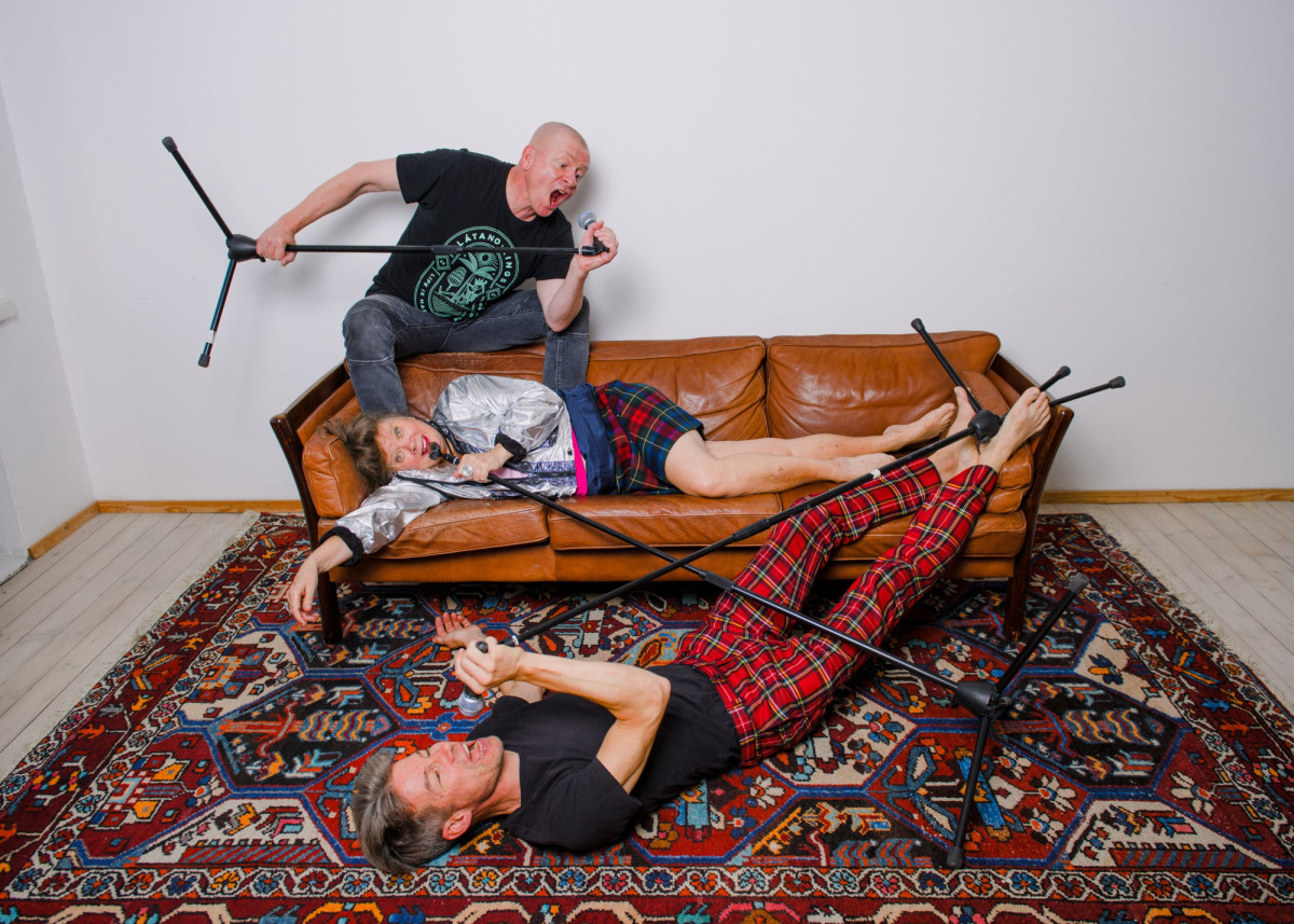 Die Performer*innen, mit Mikrofonen auf einem Teppich und einem Sofa, in die Mikrofone schreiend