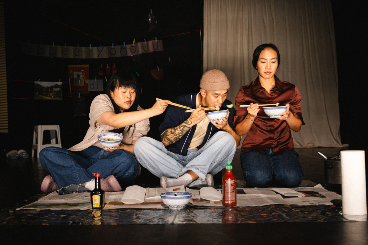 Drei Protagonist*innen sitzen auf der B&uuml;hne und essen Nudelsuppe aus wei&szlig;-blauen Schalen