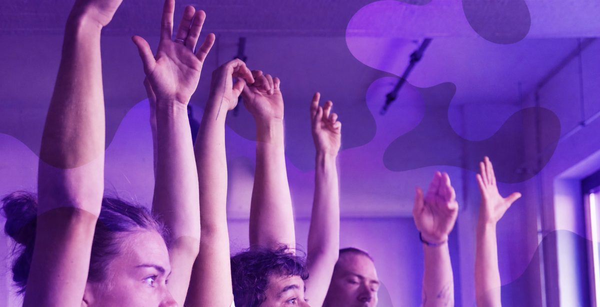 Mehrere Personen heben in einem violett beleuchteten Raum die Arme in die H&ouml;he.