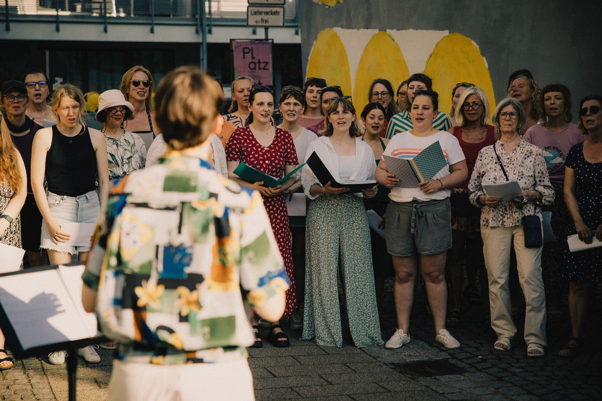Ein Chor singt drau&szlig;en auf einem Platz unter Anleitung einer Chorleitung