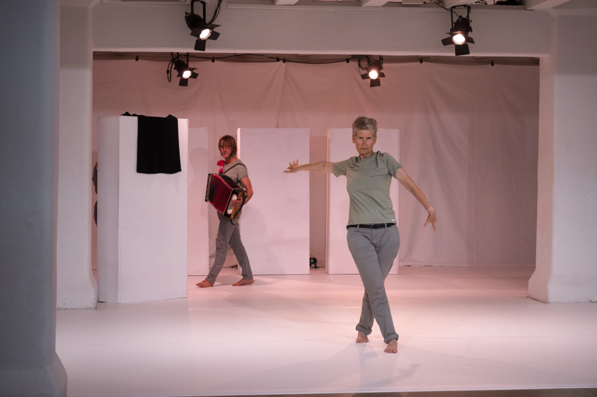 Eine wei&szlig;e B&uuml;hne, im Vordergrund eine Performer*In mit ausgebreiteten Armen, im Hintergrund eine Akteur*In mit Akkordeon