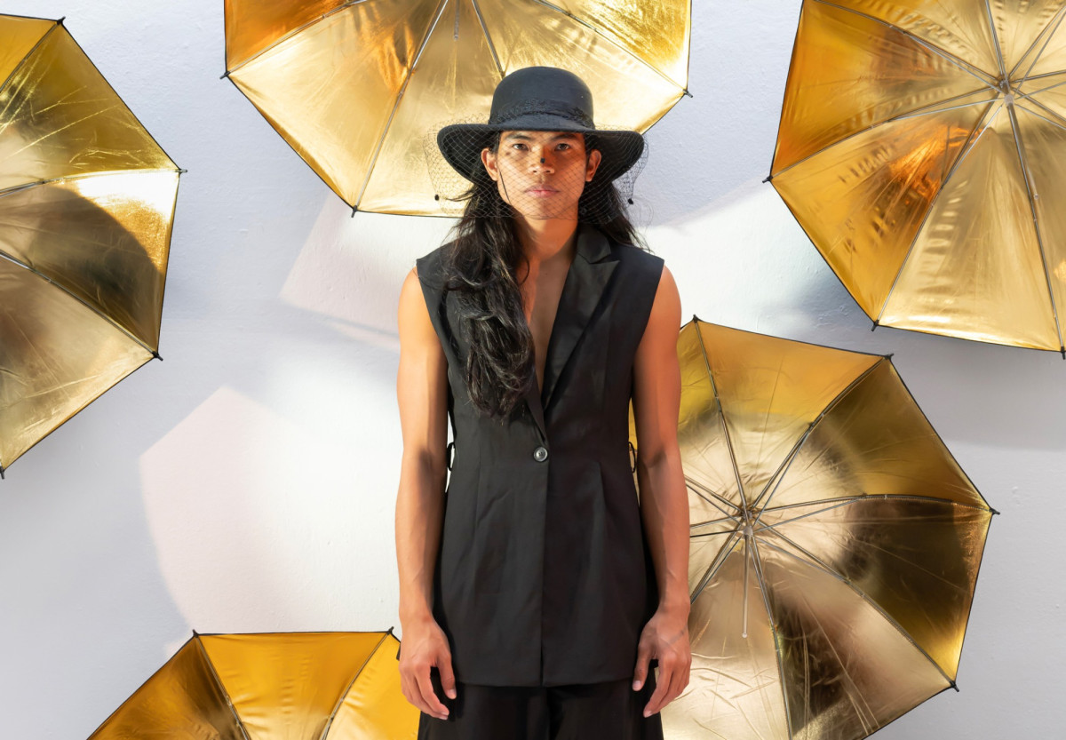 Der T&auml;nzer Jed Nagales in einem schwarzen &auml;rmellosen Anzug, im Hintergrund aufgespannte goldene Regenschirme