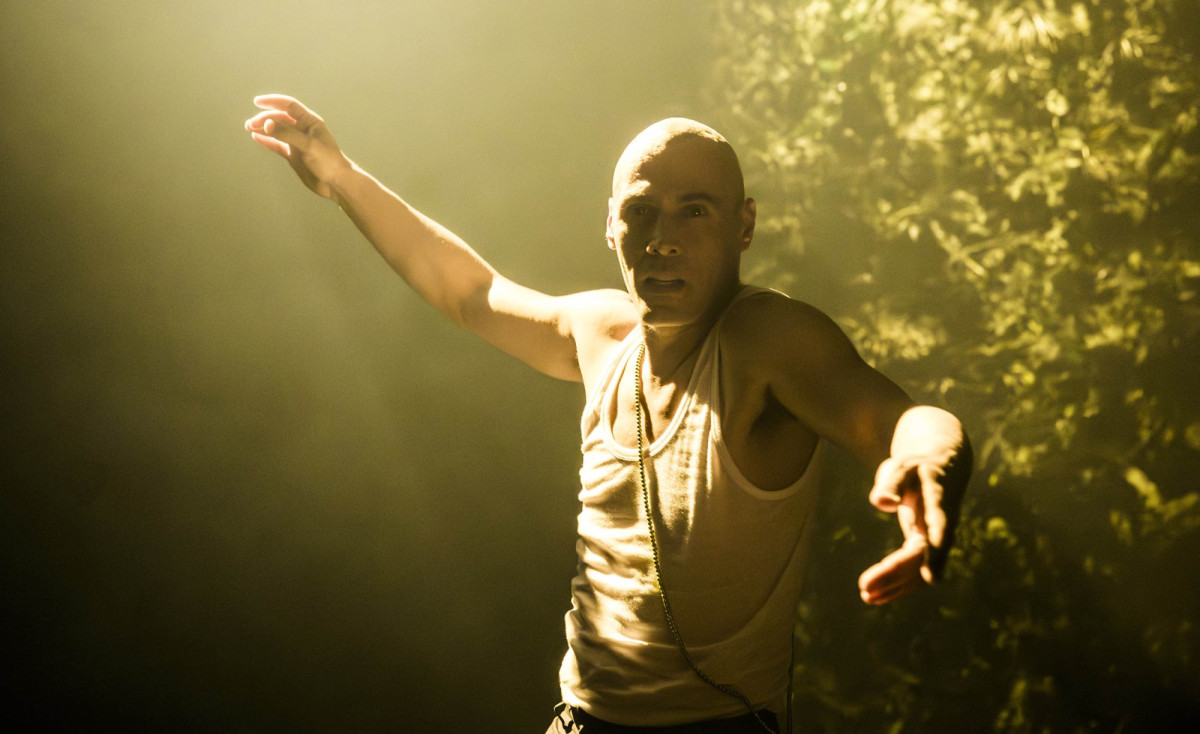 Performer mit zur Seite gestreckten Armen vor einer Wand aus umnebelten Pflanzenmaterial