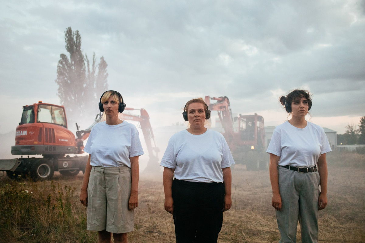 Three Pefromers are standing in front of excavators on a wasteland