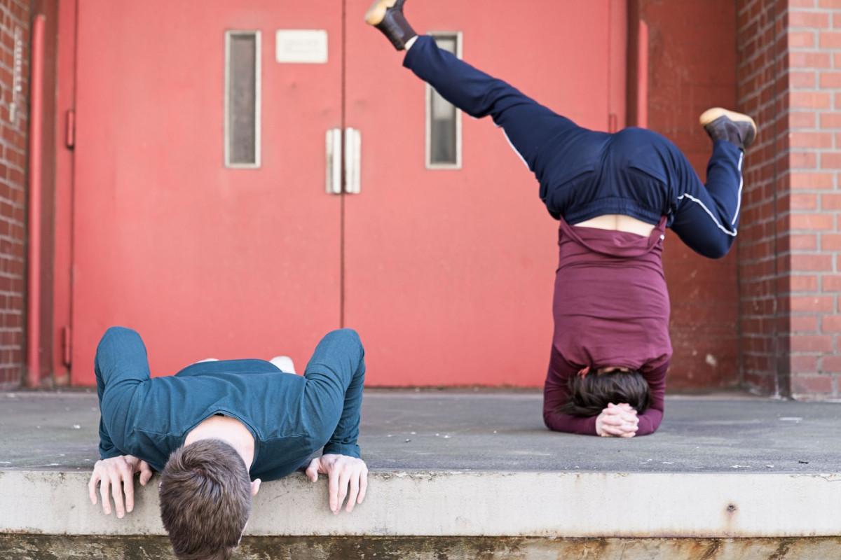 Zwei Personen agieren vor einem Hauseingang mit Laderampe. Eine Person liegt auf dem Boden mit aufgest&uuml;tzten Armen nd schaut nach unten. Die andere Person macht einen Kopfstand, die Beine zur Seite gestreckt.